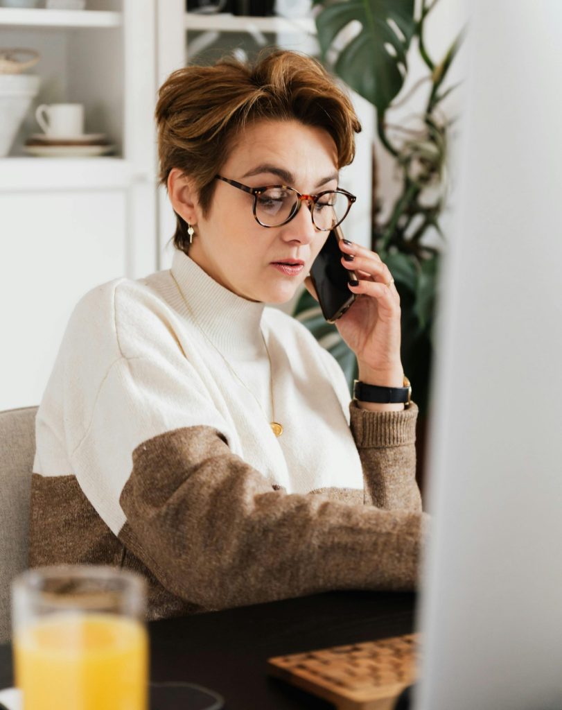Strict female director talking on mobile phone while working on computer at desk with orange juice in homelike office